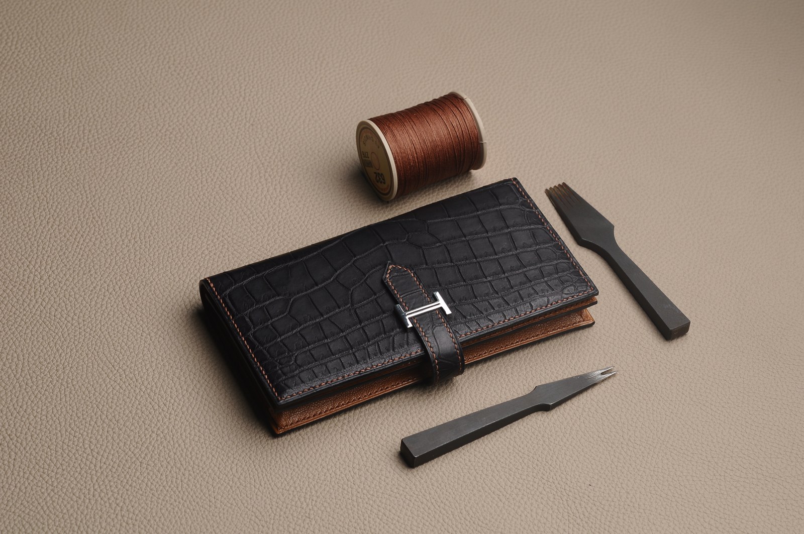 Closed black crocodile leather wallet with a metal logo, thread and hand tools